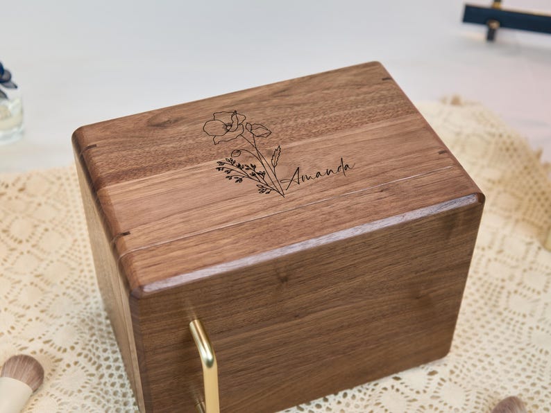May include: A rectangular wooden box with a dark brown grain finish, featuring a gold-colored metal handle on the front. The top surface is engraved with a black line-art illustration of a poppy flower and the name Amanda in script font.