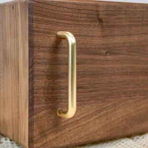 May include: A rectangular dark brown wooden box featuring a smooth, polished grain finish and a single vertical brushed gold metal handle attached to the front panel. The box has rounded edges and a solid construction with a visible wood texture.