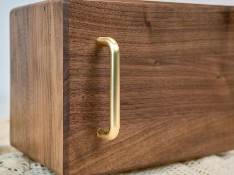 May include: A rectangular dark brown wooden box featuring a smooth, polished grain finish and a single vertical brushed gold metal handle attached to the front panel. The box has rounded edges and a solid construction with a visible wood texture.