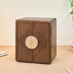 May include: A dark brown wooden jewelry box with a square silhouette and double doors. The front features a central circular gold-toned metal latch with a hammered texture, set against a carved four-lobed decorative panel on the wood surface.