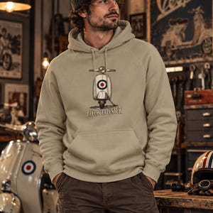 Könnte beinhalten: Ein beigefarbener Kapuzenpullover mit einer Grafik eines Oldtimer-Rollers, der vorne ein rot-weiß-blaues kreisförmiges Emblem trägt. Unter dem Roller ist der Text BLECHGEFLÜSTER in einer fetten, dunklen Schrift gedruckt. Der Hoodie hat lange Ärmel, eine Kängurutasche und passende Kordelzüge.