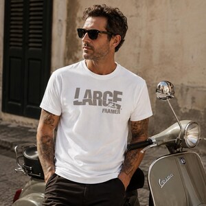 Könnte beinhalten: Weißes T-Shirt mit Rundhalsausschnitt und dem Text "LARGE FRAMER" in Grau. Das T-Shirt wird von einer Person mit Sonnenbrille getragen, die neben einem Vintage-Roller steht. Der Roller ist hellgrün mit dem Wort "Vespa" an der Seite.