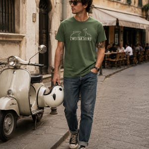 May include: A short-sleeved olive green t-shirt featuring a white line drawing of a scooter and the text ZWEITAKTKIND printed on the chest. The shirt is paired with blue denim jeans, black sneakers, and a white helmet held in one hand.