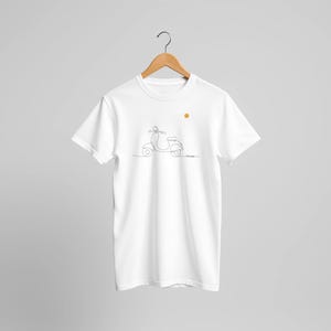 Op de afbeelding: Een wit T-shirt met korte mouwen en een minimalistische zwarte lijntekening van een scooter op de borst. Boven de scooter staat een kleine, effen oranje cirkel die de zon voorstelt. Het T-shirt hangt aan een houten kledinghanger.