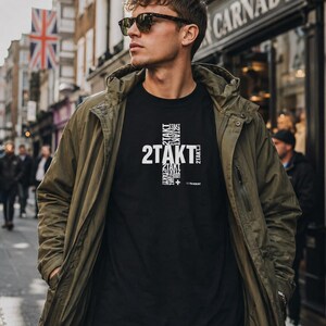 Könnte beinhalten: Ein schwarzes T-Shirt mit einem weißen Grafikdesign, bei dem der Text 2TAKT in Kreuzform angeordnet ist, getragen unter einer olivgrünen Kapuzenjacke. Das Shirt enthält kleinere, wiederholte 2TAKT-Textelemente und ein kleines Pluszeichen.