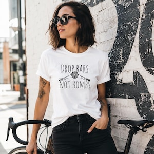 Könnte beinhalten: Ein weißes kurzärmeliges T-Shirt mit einer schwarzen Grafik von Fahrradlenkern und einem Scheinwerfer sowie dem Text "DROP BARS NOT BOMBS", der in einer einfachen serifenlosen Schrift mittig auf der Brust gedruckt ist.