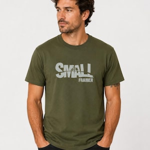 Könnte beinhalten: Olivgrünes T-Shirt mit dem Text "SMALL FRAMER" in hellgrauer, leicht verwaschener Schrift. Das kurzärmelige Shirt wird mit schwarzen Jeans kombiniert. Das Model trägt das T-Shirt.