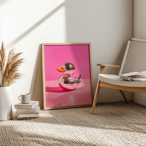 Puede incluir: Un pato de goma plateado con forma de bola de discoteca sobre un fondo rosa. El pato est&aacute; enmarcado en un marco de madera.