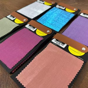 May include: A collection of six rectangular fabric swatches mounted on black cards with brown leather-like tabs secured by silver rivets. The swatches feature various colors including silver, blue, light green, magenta, purple, and peach, each with pinked edges.