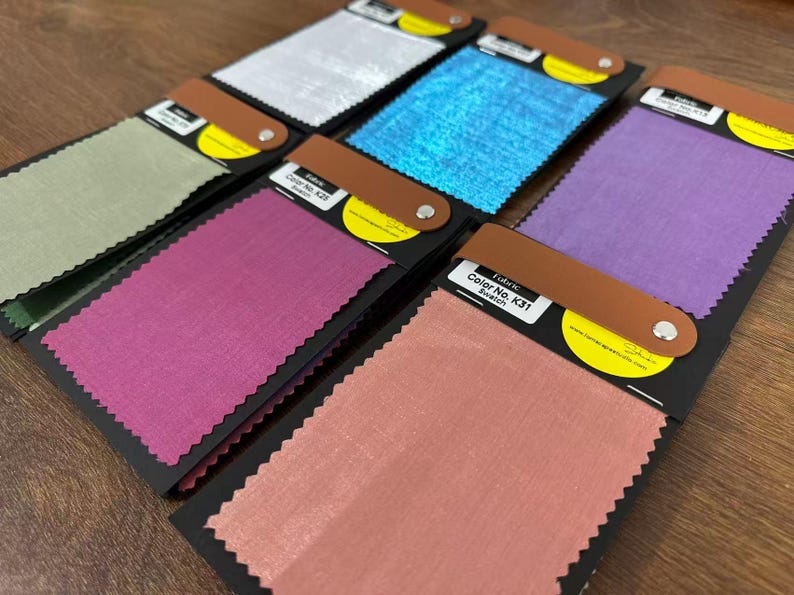 May include: A collection of six rectangular fabric swatches mounted on black cards with brown leather-like tabs secured by silver rivets. The swatches feature various colors including silver, blue, light green, magenta, purple, and peach, each with pinked edges.