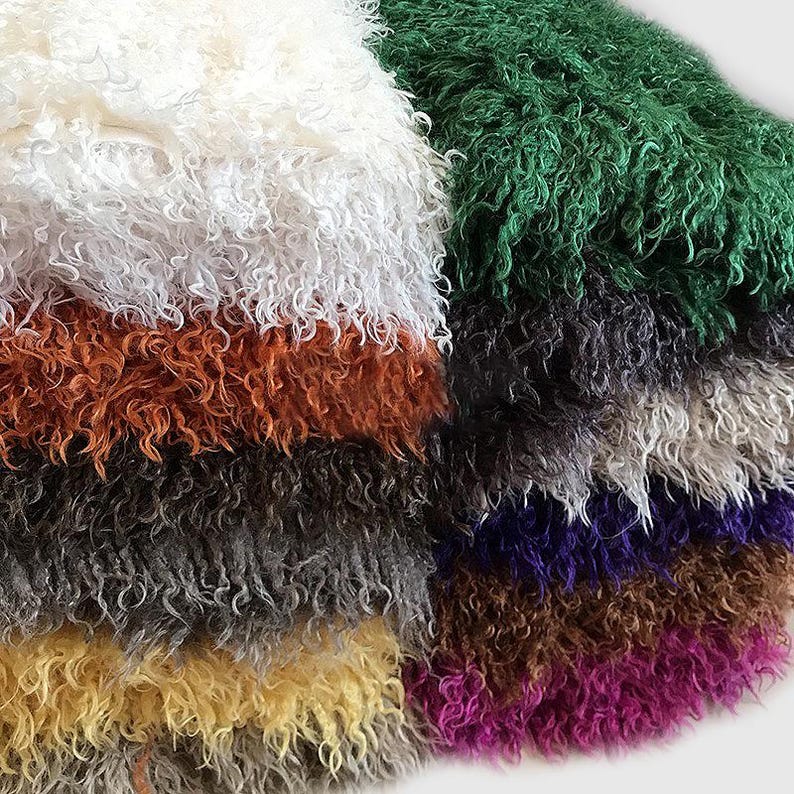 May include: A stack of folded faux fur fabric pieces in various colors including white, orange, dark gray, yellow, green, purple, and magenta. Each piece features a long, curly, shaggy texture that creates a dense and voluminous appearance.
