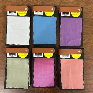 May include: Six rectangular fabric swatches arranged in two rows of three. Each swatch is attached to a black card with a tan leather-like strap and a silver rivet. The fabric colors are white, blue, purple, light green, magenta, and coral pink.