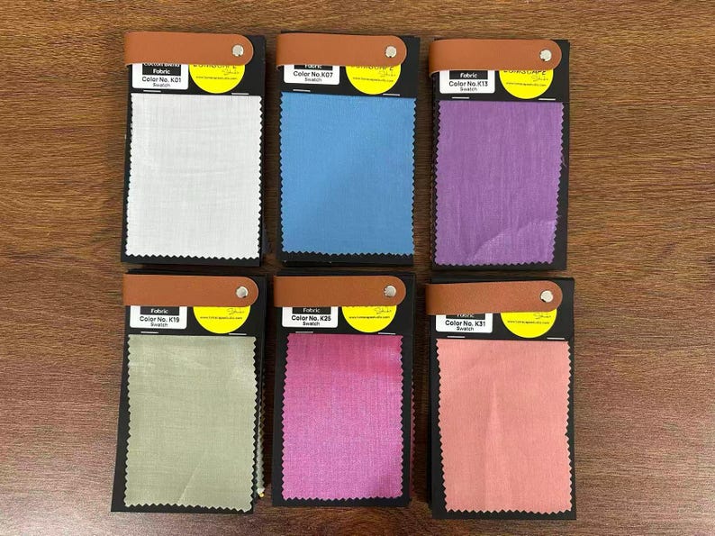 May include: Six rectangular fabric swatches arranged in two rows of three. Each swatch is attached to a black card with a tan leather-like strap and a silver rivet. The fabric colors are white, blue, purple, light green, magenta, and coral pink.