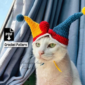 Peut inclure: Un chat blanc portant un chapeau de bouffon crocheté coloré. Le chapeau est jaune, rouge et bleu avec une bordure blanche et des pompons. L'image comprend le texte "Crochet Pattern" et le logo "PDF". Le chat a les yeux verts.