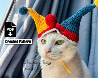 Könnte beinhalten: Eine weiße Katze mit einem bunten, gehäkelten Narrenhut. Der Hut ist gelb, rot und blau mit weißem Besatz und Bommeln. Das Bild enthält den Text "Crochet Pattern" und das Logo "PDF". Die Katze hat grüne Augen.