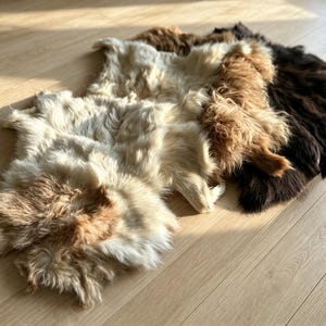 Pode incluir: Três tapetes de pele de ovelha com texturas de pelo longo e felpudo. Os tapetes estão dispostos em camadas, apresentando variações de cores distintas, incluindo um branco-creme, um castanho-claro quente e um castanho-escuro profundo.