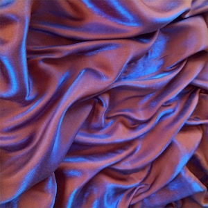 May include: A smooth, lustrous fabric featuring a deep plum or dark purple hue with vibrant blue highlights. The material is draped in soft, flowing folds that create a dynamic interplay of light and shadow across the surface of the textile.