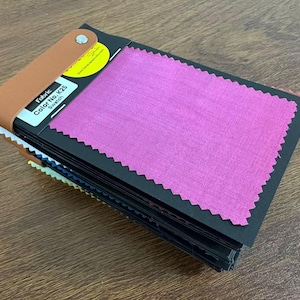 May include: A fabric swatch book featuring a stack of black cards with pinked edges. The top card displays a vibrant magenta fabric sample and a white label with the text Fabric Color No. K25 Swatch. A tan leather strap secures the stack with a silver rivet.