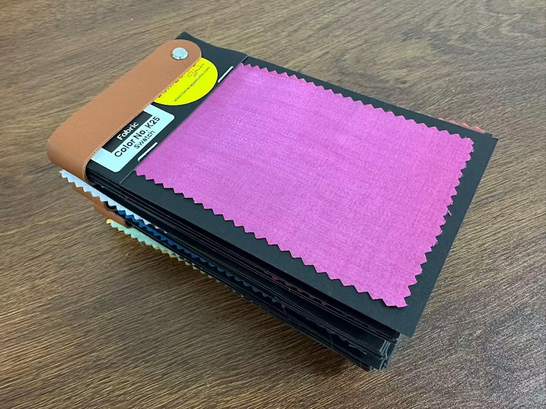 May include: A fabric swatch book featuring a stack of black cards with pinked edges. The top card displays a vibrant magenta fabric sample and a white label with the text Fabric Color No. K25 Swatch. A tan leather strap secures the stack with a silver rivet.