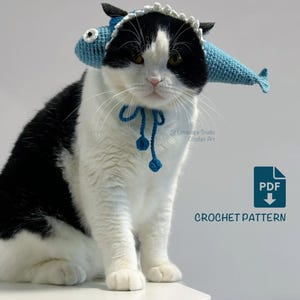 May include: A black and white cat wearing a blue crocheted fish hat with a matching blue tie. The hat has a white scalloped trim and the fish have black fins. The image includes the text "CROCHET PATTERN" and a PDF icon.