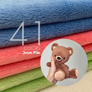 May include: A stack of soft, fuzzy fabric in blue, coral, and light green with a 3mm pile. The text 41 COLORS 3mm Pile is printed on the fabric. A circular inset shows a brown plush teddy bear with light tan accents on the ears and feet.