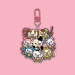 May include: A pink keychain featuring a cluster of cartoon characters with various designs, including a cat, bunny, and bear. The characters are in pastel colors with star-shaped accents. The keychain has a pink clasp and a pink background.