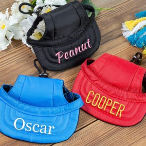 May include: Three dog harnesses with adjustable straps and a hat-like brim. The harnesses are blue, red, and black, and each has a name embroidered on the brim: Oscar, Cooper, and Peanut.