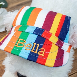 May include: A white dog wearing a colorful striped hat with the name "Bella" embroidered on the front.