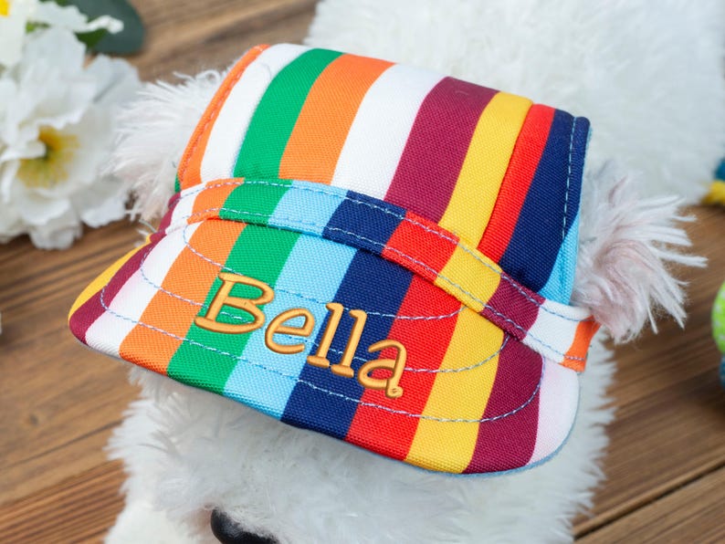 May include: A white dog wearing a colorful striped hat with the name "Bella" embroidered on the front.