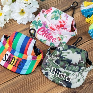 May include: Three dog hats with personalized names. The hats are in a floral print, a rainbow stripe pattern, and a camouflage print. The names on the hats are Chloe, Sadie, and Buster.