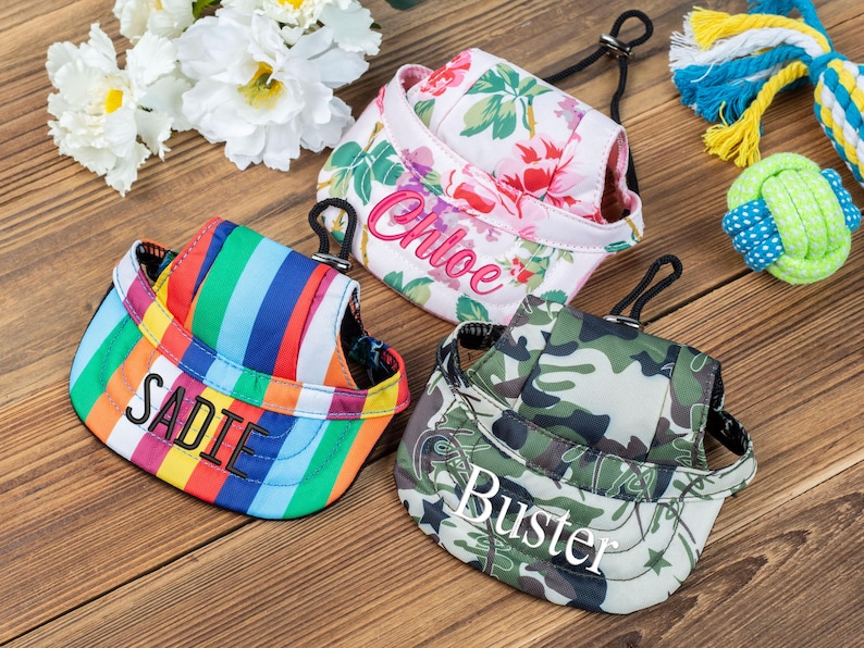 May include: Three dog hats with personalized names. The hats are in a floral print, a rainbow stripe pattern, and a camouflage print. The names on the hats are Chloe, Sadie, and Buster.