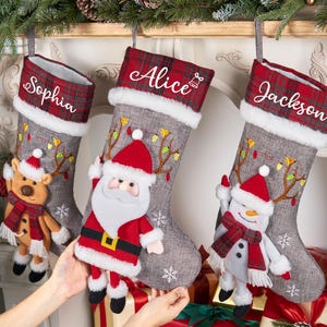 以下が含まれることがあります： 赤と白のチェック柄の袖口に「Sophia」、「Alice」、「Jackson」と刺繍された3つのクリスマスストッキング。各グレーのストッキングには白いフェイクファーのトリムがあり、トナカイ、サンタ、雪だるまで飾られています。暖炉に飾られています。