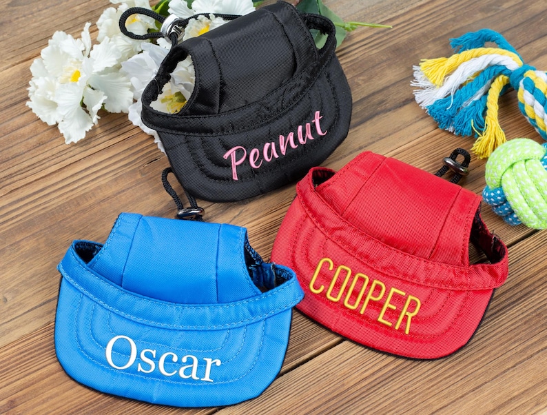 May include: Three dog harnesses with adjustable straps and a hat-like brim. The harnesses are blue, red, and black, and each has a name embroidered on the brim: Oscar, Cooper, and Peanut.