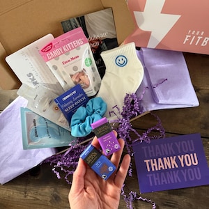 Peut inclure: Une boîte en carton ouverte contenant divers articles, dont une chaussette blanche avec un visage souriant, un chouchou bleu et de petites barres de chocolat. On y trouve également une boîte rose, du papier de soie violet et une carte de remerciement.