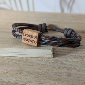 Könnte beinhalten: Ein Armband mit Camouflage-Muster und einem rechteckigen Mittelstück aus Holz. Das Holzstück ist mit den Koordinaten "54°09'31"N 10°80'40"O" graviert. Das Armband besteht aus einem strapazierfähigen, gewebten Material.