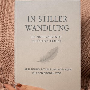 Könnte beinhalten: Ein Buch mit dem Titel "IN STILLER WANDLUNG" wird gehalten. Das Buchcover ist hellgrau mit dunkelgrauem Text und einer Federillustration. Der Text enthält Sätze wie "EIN MODERNER WEG DURCH DIE TRAUER" und "BEGLEITUNG, RITUALE UND HOFFNUNG".