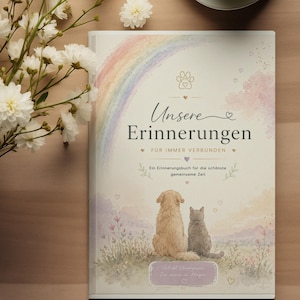 May include: A hardback memory book with the German text "Unsere Erinnerungen" (Our Memories) featuring a rainbow, paw print, and illustrations of a dog and cat. A cup of tea and white flowers are also in the image.