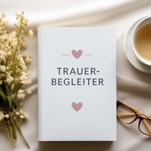 Könnte beinhalten: Ein weißes Buch mit dem Text "TRAUER-BEGLEITER" und zwei rosa Herzen. Eine Tasse Tee, eine Brille und ein Strauß weißer Blumen sind um das Buch auf einer cremefarbenen Oberfläche angeordnet.