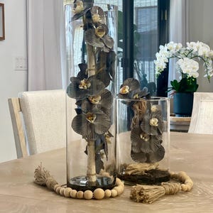 Peut inclure: Deux vases cylindriques en verre transparent contenant des orchidées noires avec des tiges en bois. Les vases sont posés sur une table en bois avec une guirlande de perles en bois et un pompon en jute.