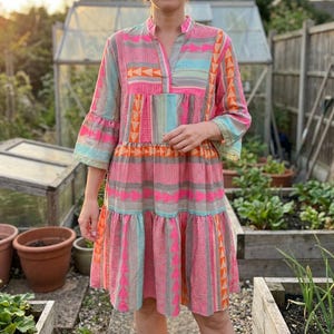 Könnte beinhalten: Ein gestuftes knielanges Kleid mit einem lebendigen geometrischen Muster in Neonrosa, leuchtendem Orange, Hellblau und Grau. Das Kleidungsstück hat dreiviertellange Glockenärmel, einen V-Ausschnitt und eine lockere, fließende Silhouette mit Querstreifen.