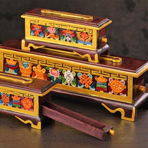 Può includere: Tre scatole rettangolari in legno per incenso, color marrone scuro con bordi dipinti in oro. Ogni scatola presenta rilievi colorati dipinti a mano con simboli sui lati, una fessura superiore e un cassetto scorrevole per gli incensi.
