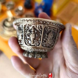 May include: A small, silver-coloured bowl with intricate carvings of four deities. The bowl is held in a hand, and the background is blurred.