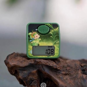 Può includere: Un contatore digitale quadrato di colore verde con un'illustrazione di un personaggio dei cartoni animati sul pannello frontale. Il dispositivo presenta un pulsante circolare verde sulla parte superiore e un piccolo schermo LCD che mostra il numero 108.