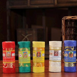 May include: Five cylindrical incense containers in red, green, yellow, white, and blue. Each features a central label with a deity illustration and text: Guru Rinpoche, Green Tara, Zambala, Chenrezig, and Medicine Buddha, with gold-toned patterns.