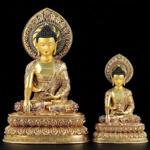 May include: Two gold Buddha statues in a seated meditation pose. The larger statue has a detailed halo with turquoise accents. Both statues are adorned with intricate patterns and colourful gemstones, set against a black background.