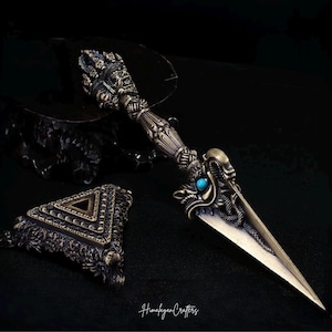 May include: A bronze-toned phurba ritual dagger featuring an intricate handle with a deity face, a central turquoise stone, and a dragon motif. Beside it lies a triangular base with ornate detailing. The text Himalayan Crafters is visible.