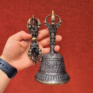 Pode incluir: Um vajra e um sino de metal em tom de bronze com um cabo ornamentado apresentando um rosto esculpido. O sino tem uma base alargada decorada com padrões em relevo intrincados e linhas verticais, enquanto o vajra tem um design simétrico.