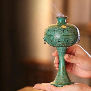 Puede incluir: Un quemador de incienso de metal color cerceta con un acabado de pátina desgastada. El recipiente presenta un patrón en relieve intrincado, una base acampanada y una tapa con pequeñas aberturas circulares. Un fino hilo de humo sale por la parte superior.