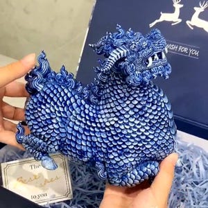 Könnte beinhalten: Eine blau-weiße Qilin-Figur aus Keramik mit komplexen Schuppenmustern, einem gehörnten Kopf und sichtbaren Zähnen. Das Fabelwesen ist mit detaillierten, wirbelnden Mähnen- und Schwanzstrukturen sowie einer kobaltblauen Glasur gestaltet.
