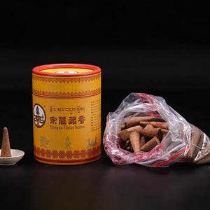 May include: A yellow cylindrical container of Dronngsar Tibetan Incense with red accents, a small white dish with a single incense cone, and a clear plastic bag filled with incense cones.